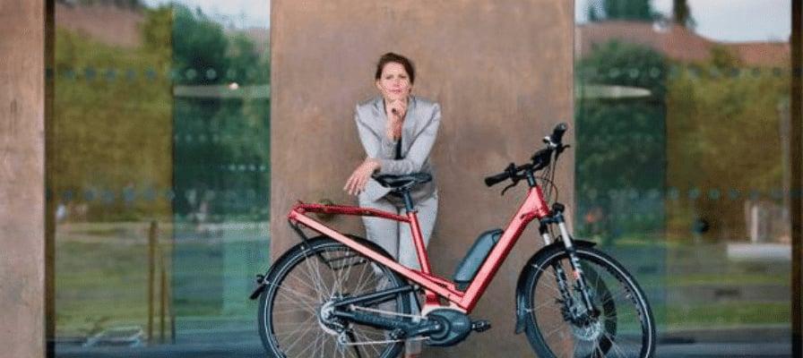 Frau in Business-Kleidung lehnt an rotem E-Bike vor Bürogebäude