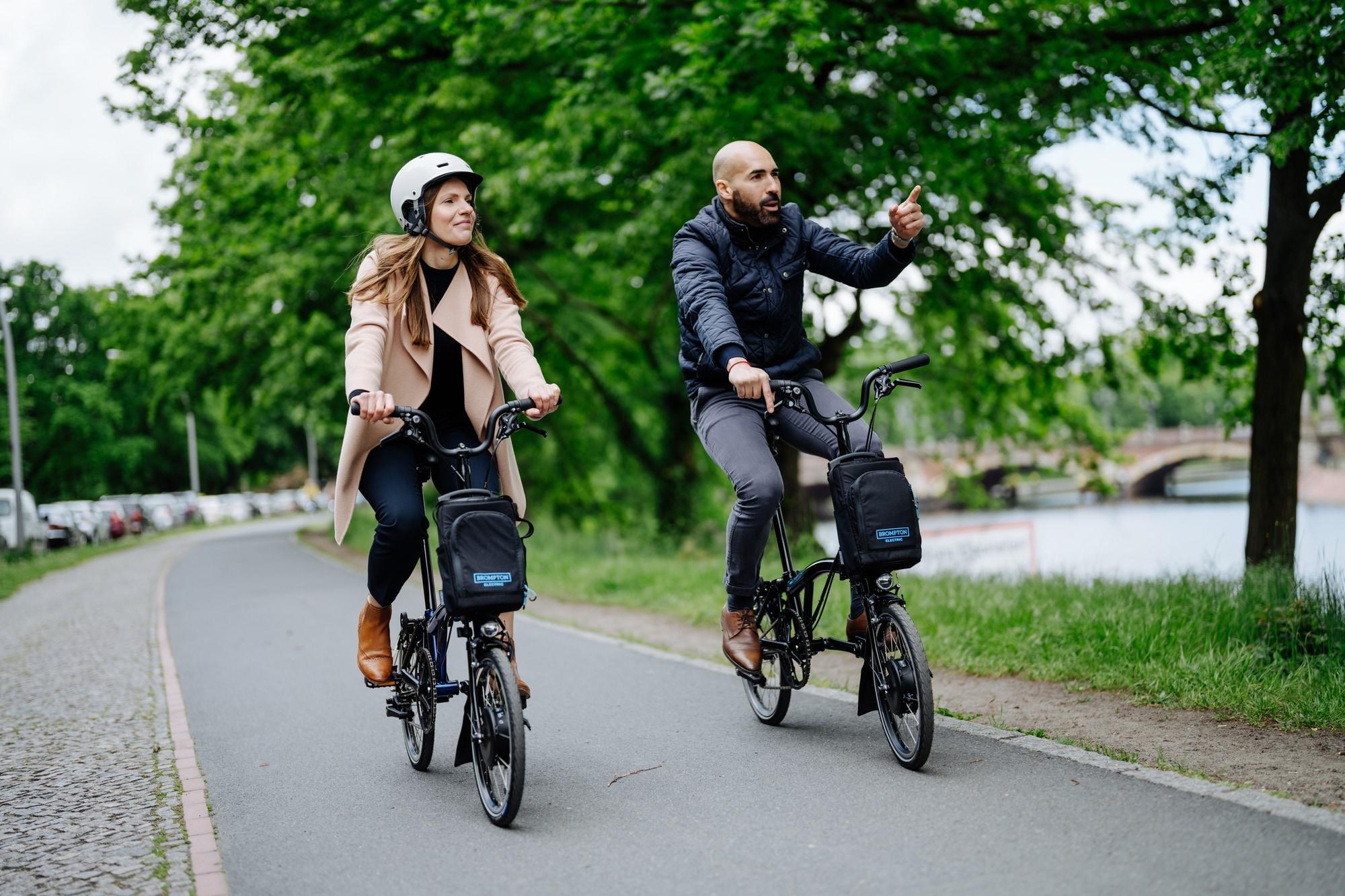 Zwei Personen fahren Brompton-Falträder entlang eines malerischen Weges. Ideal für urbane Mobilität und stilvolle A