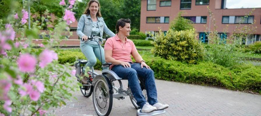 Tandem-Dreirad mit zwei Personen auf gepflastertem Weg vor Wohngebäude