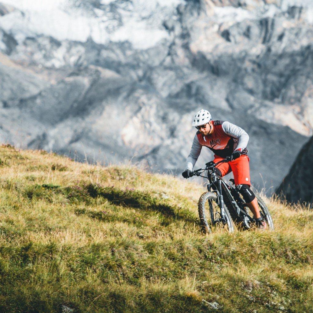 Ein Radfahrer auf einem Mountainbike fährt einen grasbewachsenen Hügel in einer alpinen Umgebung hinunter. Er trägt