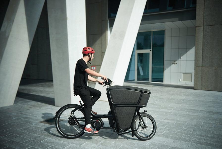 Person auf schwarzem Lastenfahrrad mit rotem Helm vor modernem Gebäude