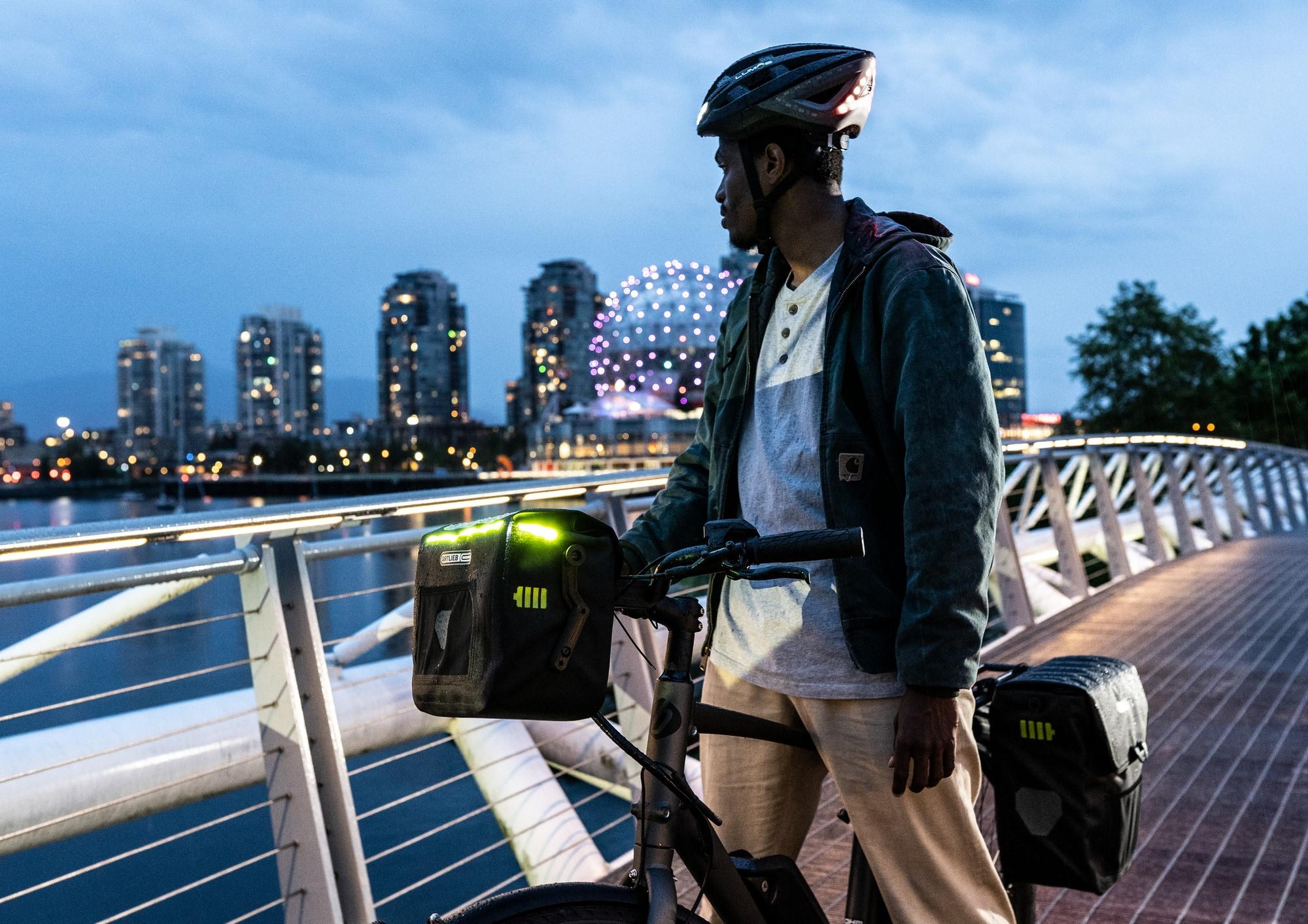 Mann auf einer Brücke mit einem VAN E-Bike und ORTLIEB Fahrradtaschen, vor einer städtischen Skyline.
