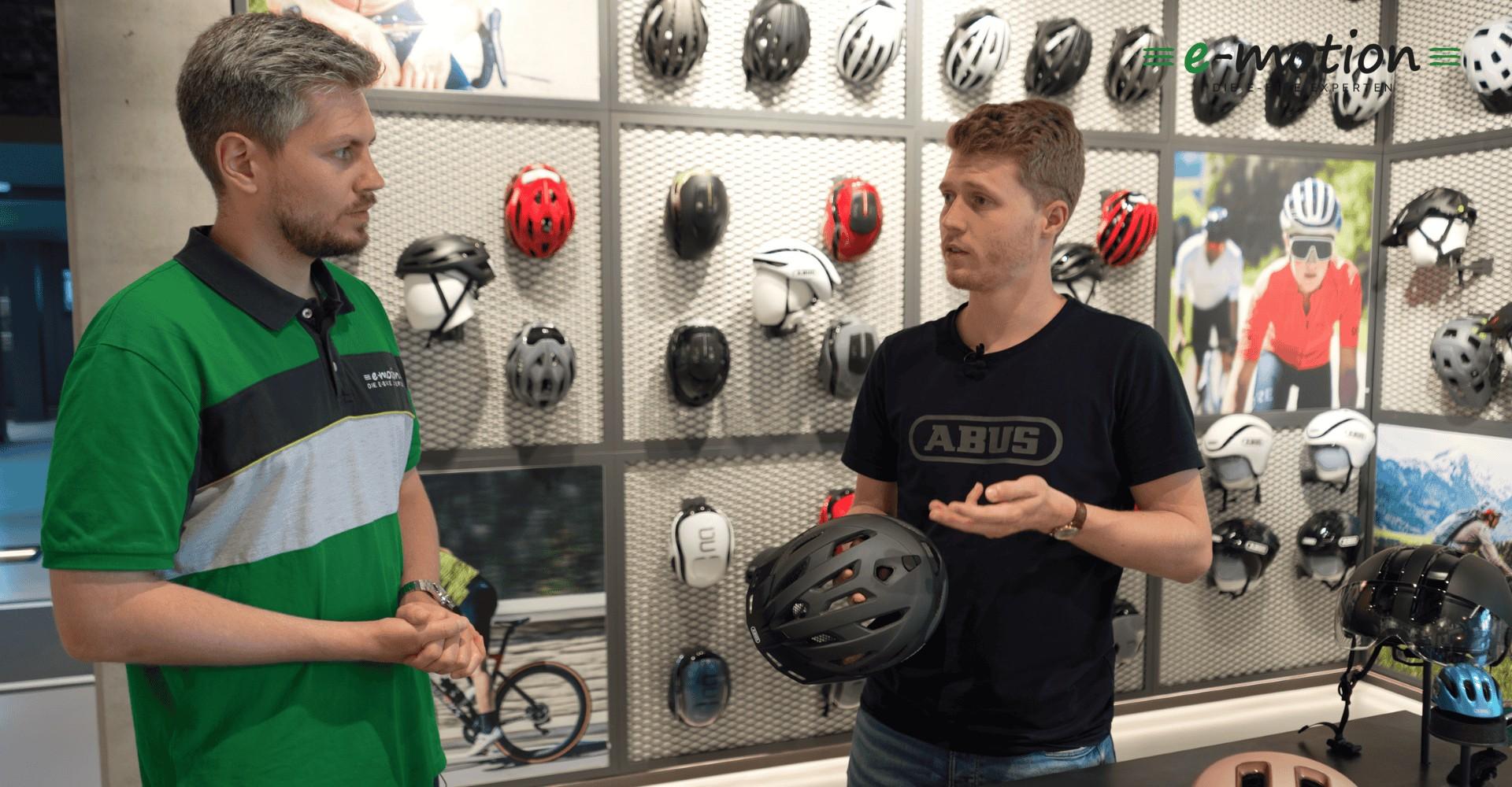 Zwei Männer stehen vor einer Wand mit Abus Helmen