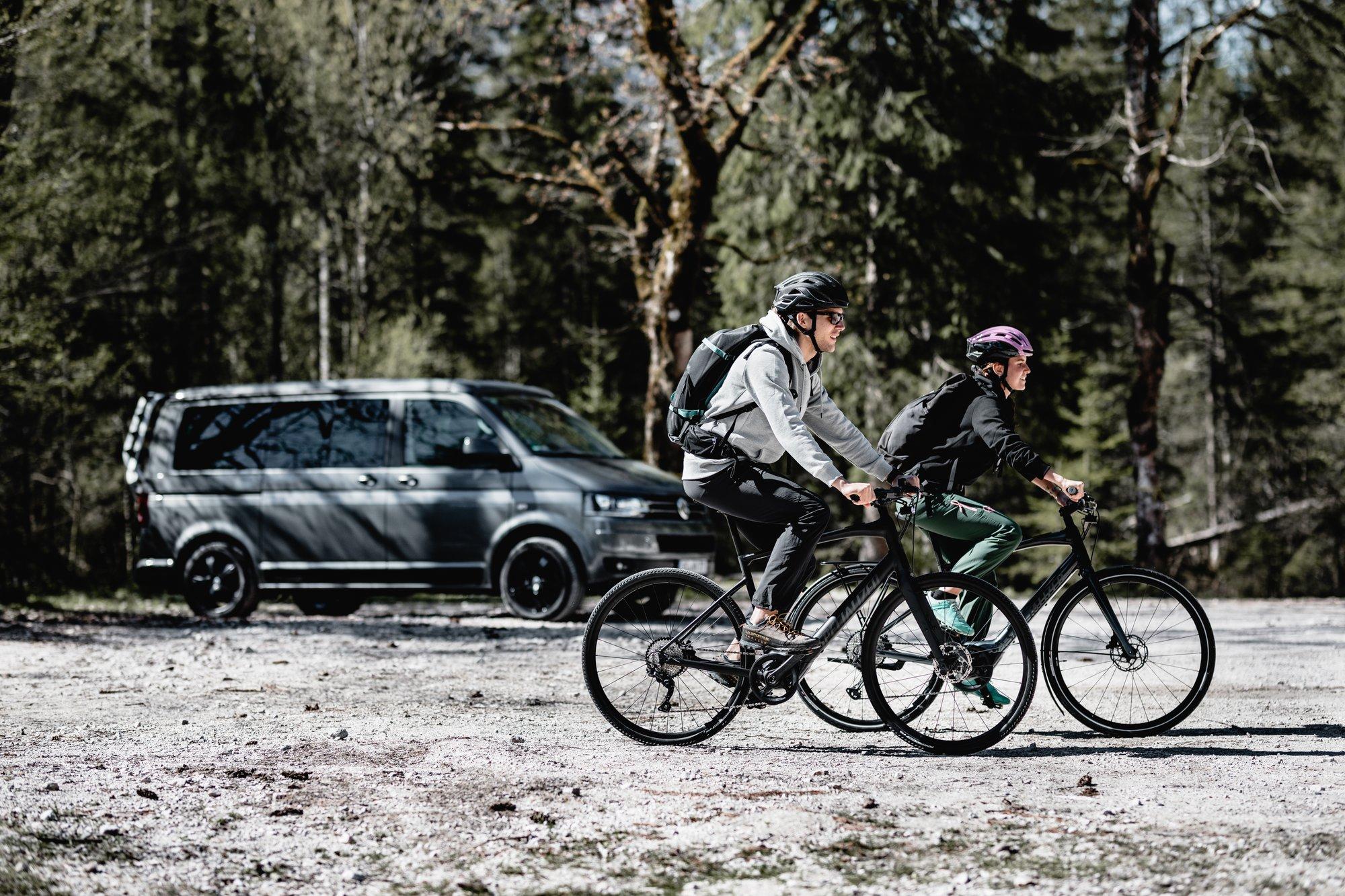 Zwei Radfahrer tragen Helme und fahren nebeneinander auf einem Fahrradweg durch einen Wald. Im Hintergrund steht ei