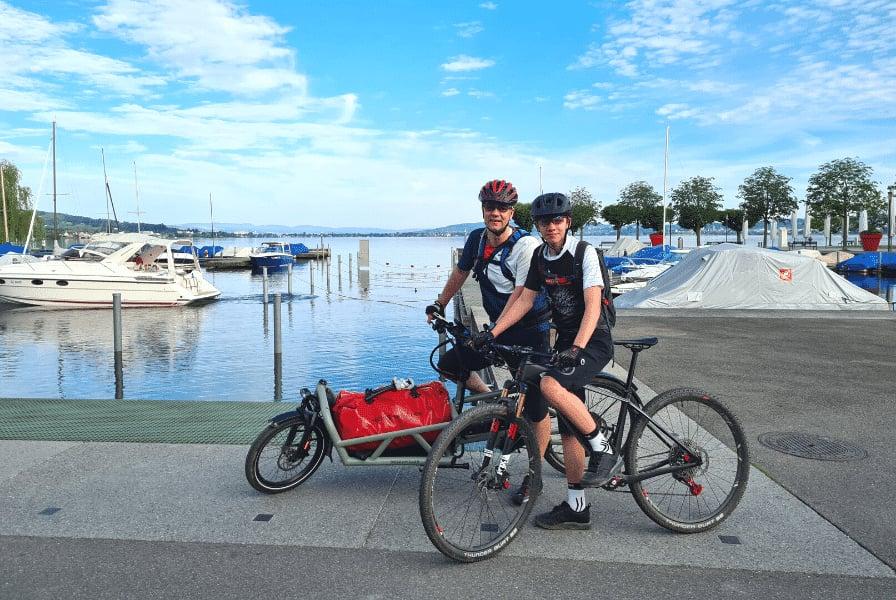 Zwei Radfahrer mit Anhänger am Hafen vor Booten und Wasser