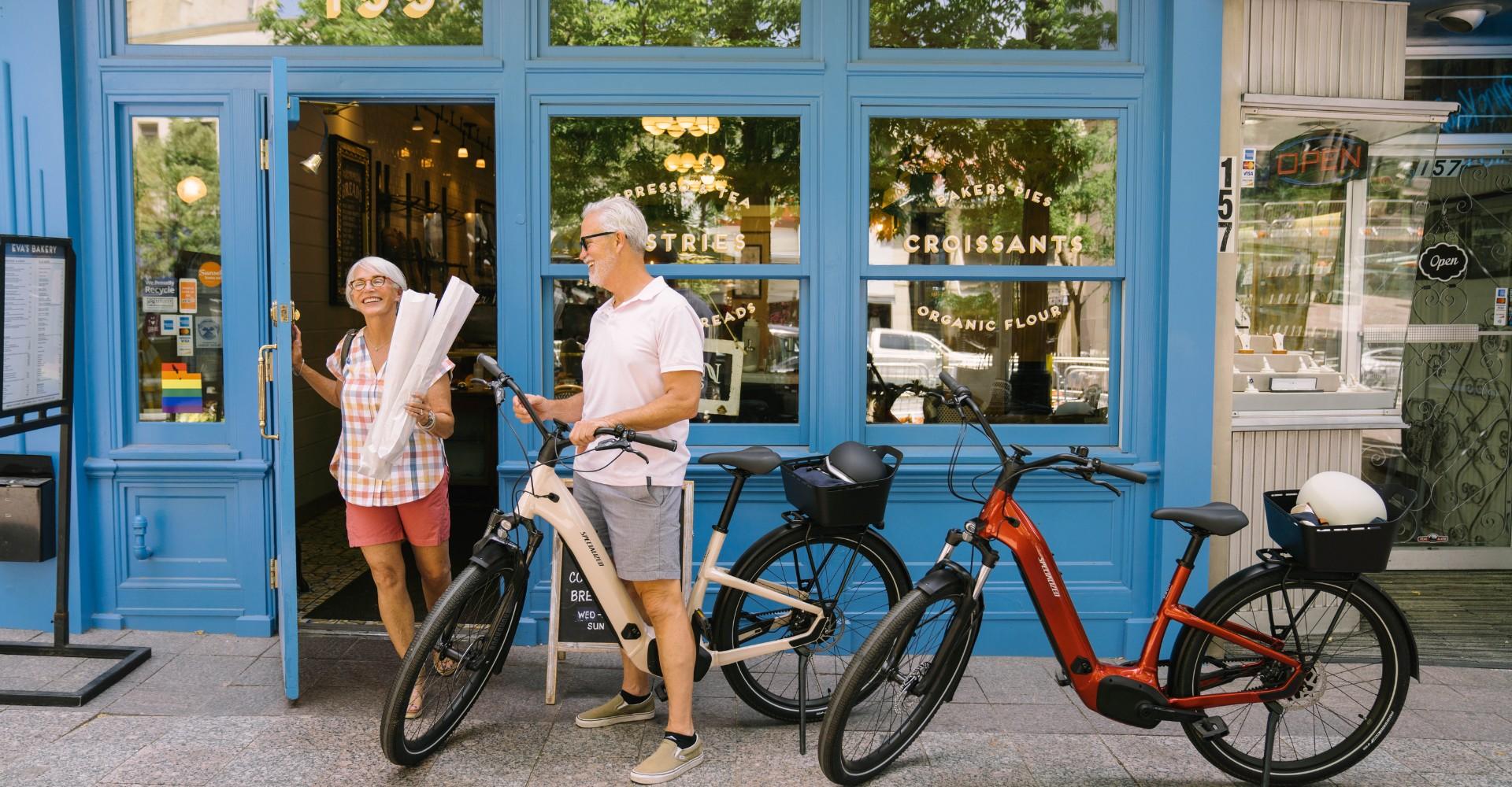 Zwei Senioren stehen vor einer Bäckerei mit ihren e-Bikes