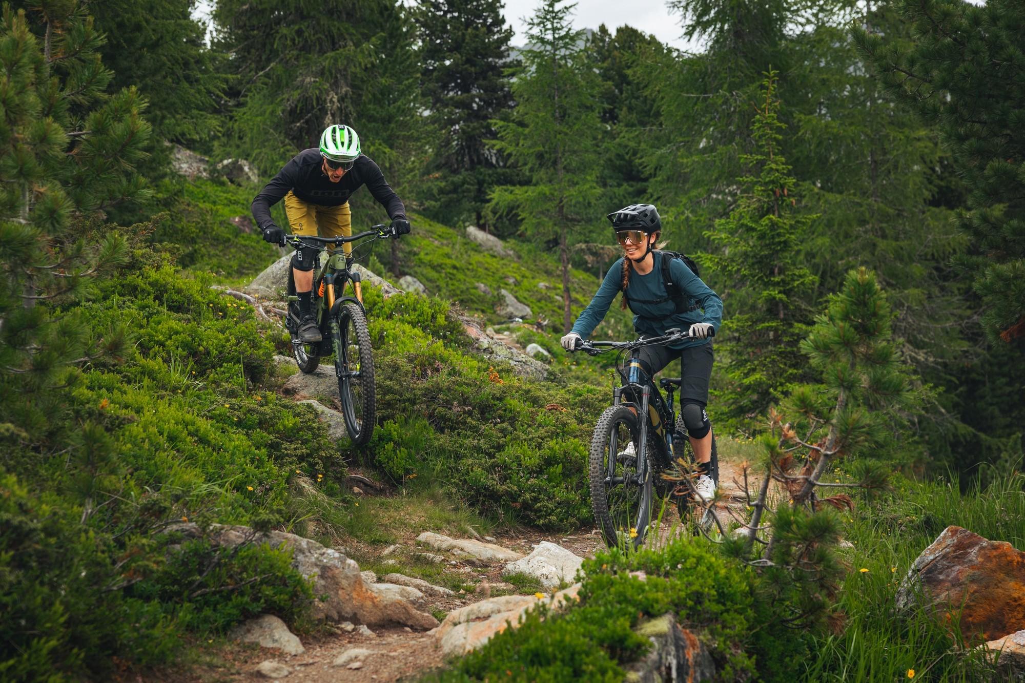 Das Bild zeigt zwei Radfahrer auf E-Bikes von R Raymon, die einen felsigen, bewaldeten Pfad entlangfahren.