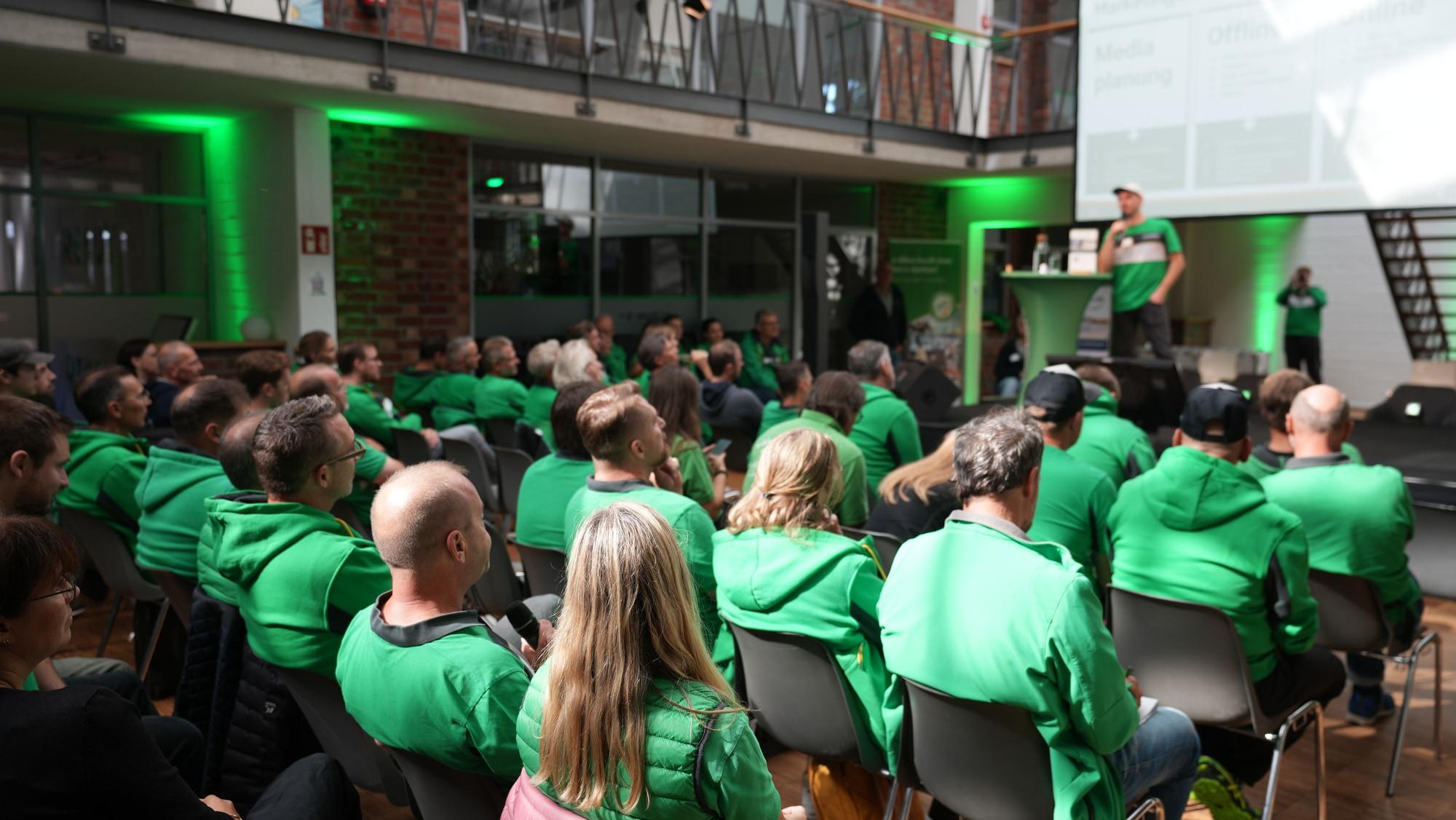 Eine große Gruppe in grüner Kleidung hört aufmerksam einem Vortrag in einem modern gestalteten Raum zu.