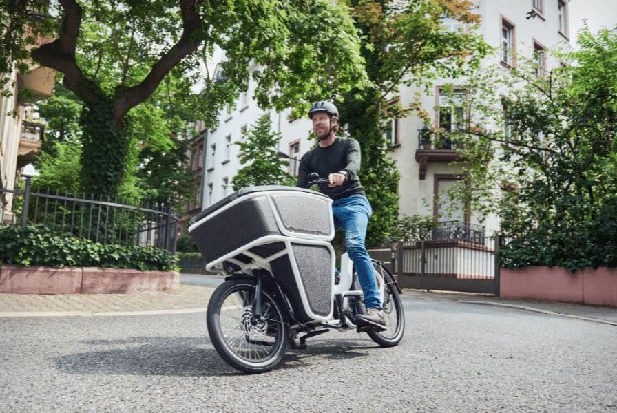 Mann fährt Lastenfahrrad auf städtischer Straße mit grüner Umgebung