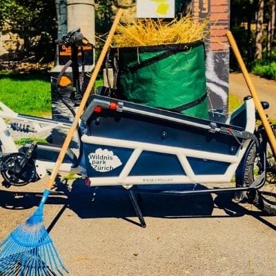 Weißes E-Bike Lastenrad vor einer Hecke mit Besen und Schaufel