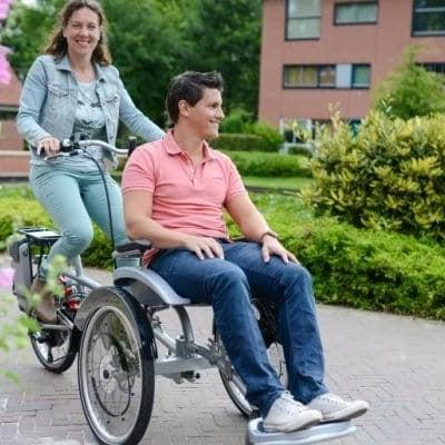 Tandem-Dreirad mit zwei Personen auf gepflastertem Weg vor Wohngebäude