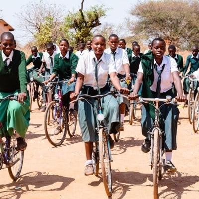 Schülerinnen fahren gemeinsam mit Fahrrädern zur Schule auf einem staubigen Weg