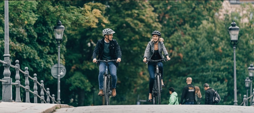 Zwei Personen fahren auf E-Bikes über eine Brücke in einer grünen Umgebung