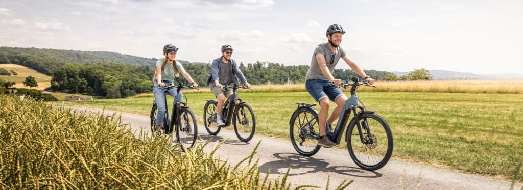 Drei Personen fahren E-Bikes auf einem Landweg bei sonnigem Wetter