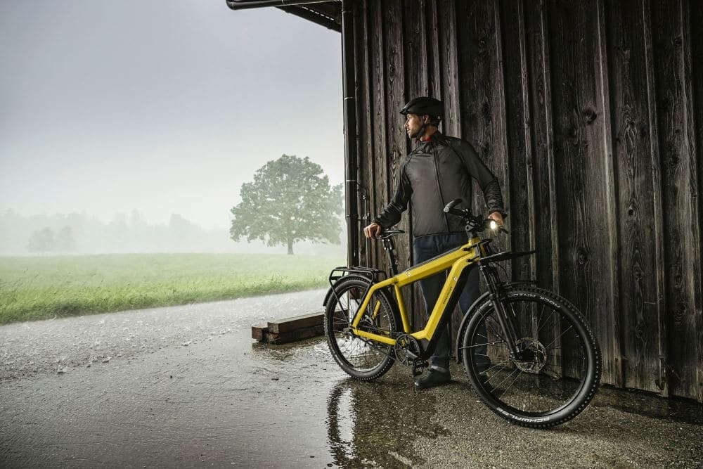 Mann mit gelbem E-Bike bei Regen vor Holzwand