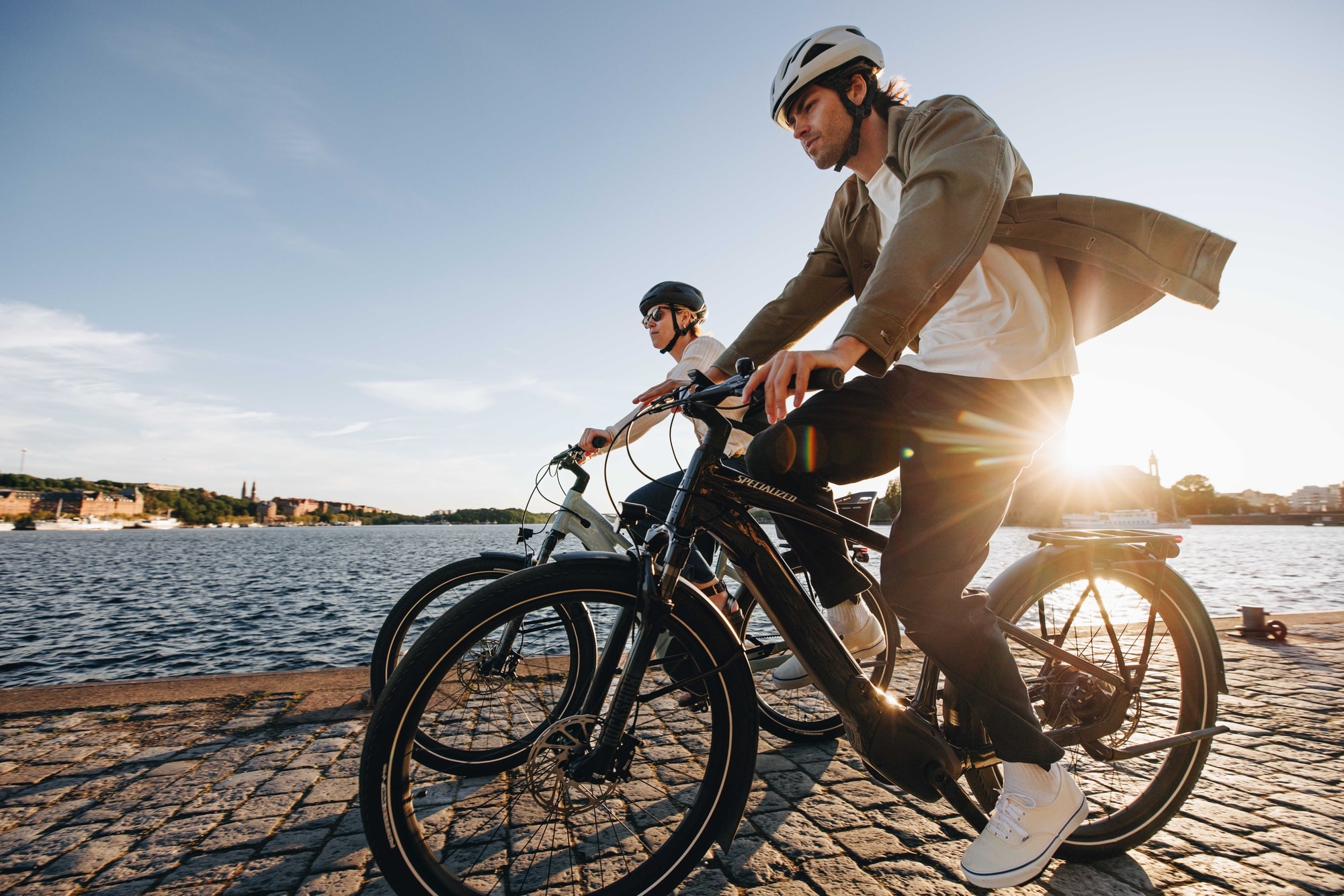 Zwei Personen fahren Specialized Vado E-Bikes entlang einer Uferpromenade bei Sonnenuntergang für ein aktives und s
