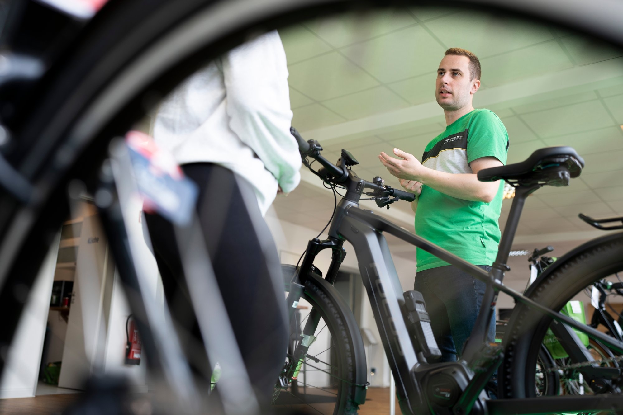 Ein Mann in einem grünen T-Shirt steht neben einem modernen E-Bike und erklärt einer anderen Person, die teilweise